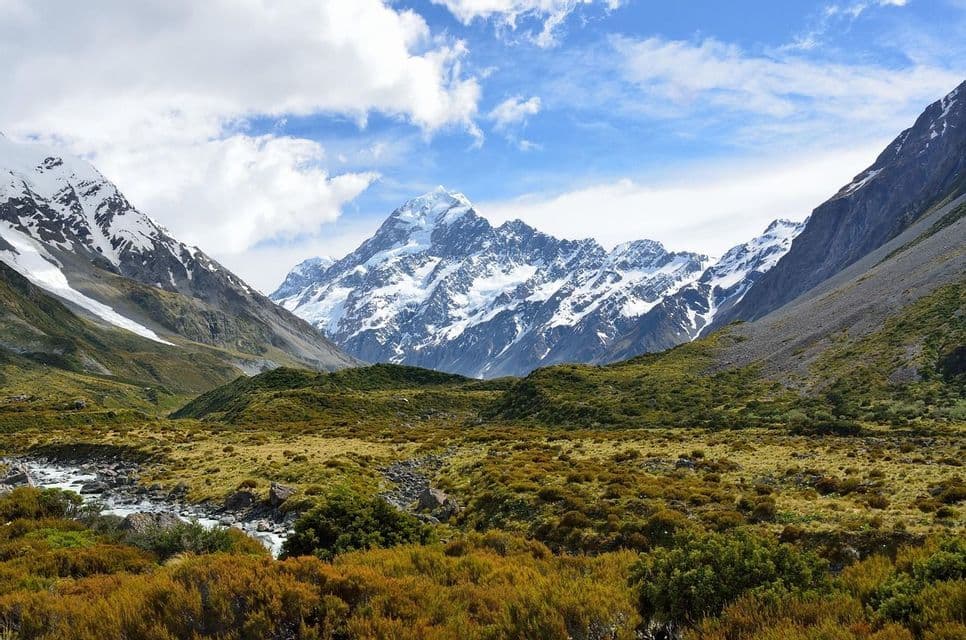 Une chaîne de montagnes enneigées s'élève au-dessus d'une vaste vallée remplie d'arbustes verts et dorés, traversée par un petit cours d'eau.