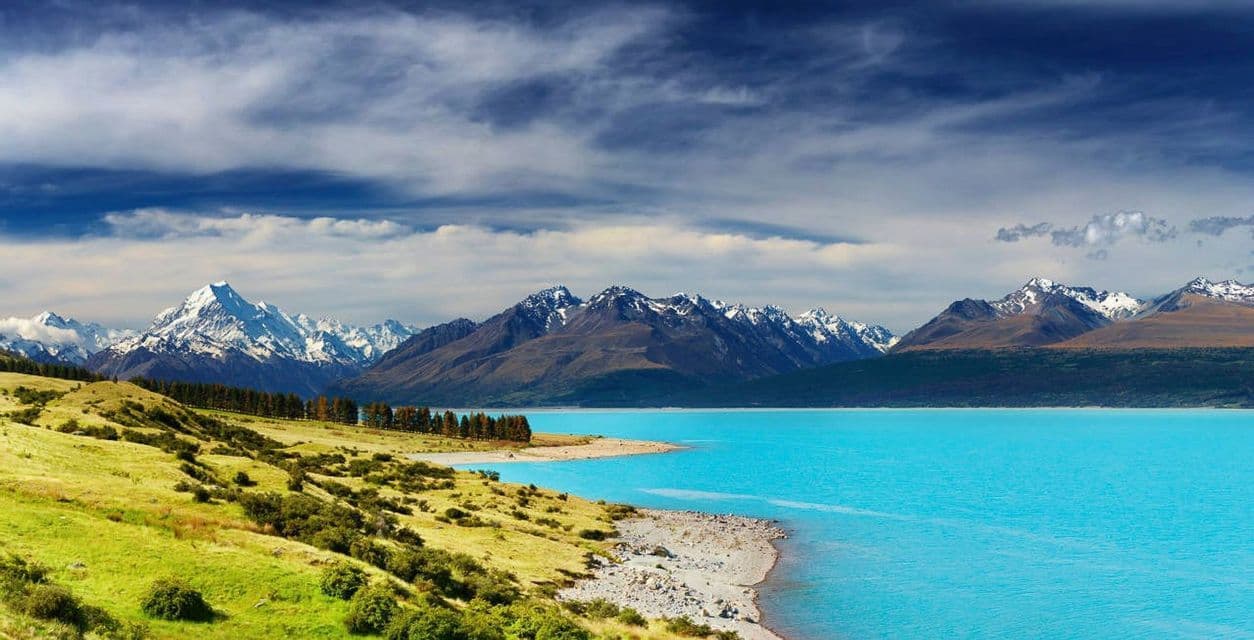 Une rive herbeuse borde un lac turquoise éclatant devant une chaîne de montagnes enneigées lointaine, sous un ciel partiellement nuageux.