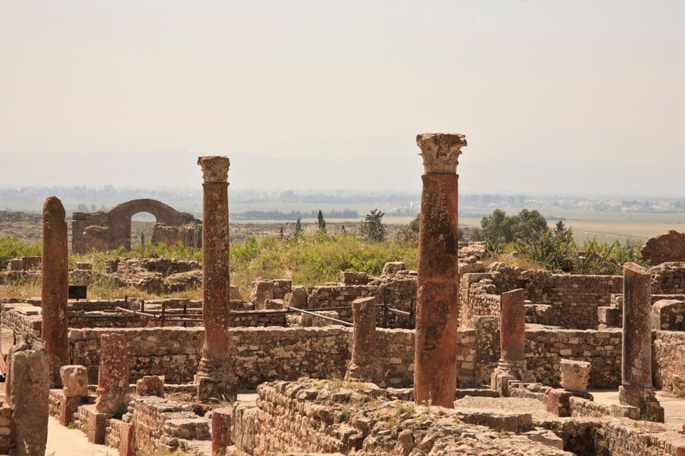 Verwitterte korinthische Säulen stehen inmitten antiker Steinruinen an einer archäologischen Stätte, mit einem dunstigen Tal im Hintergrund.