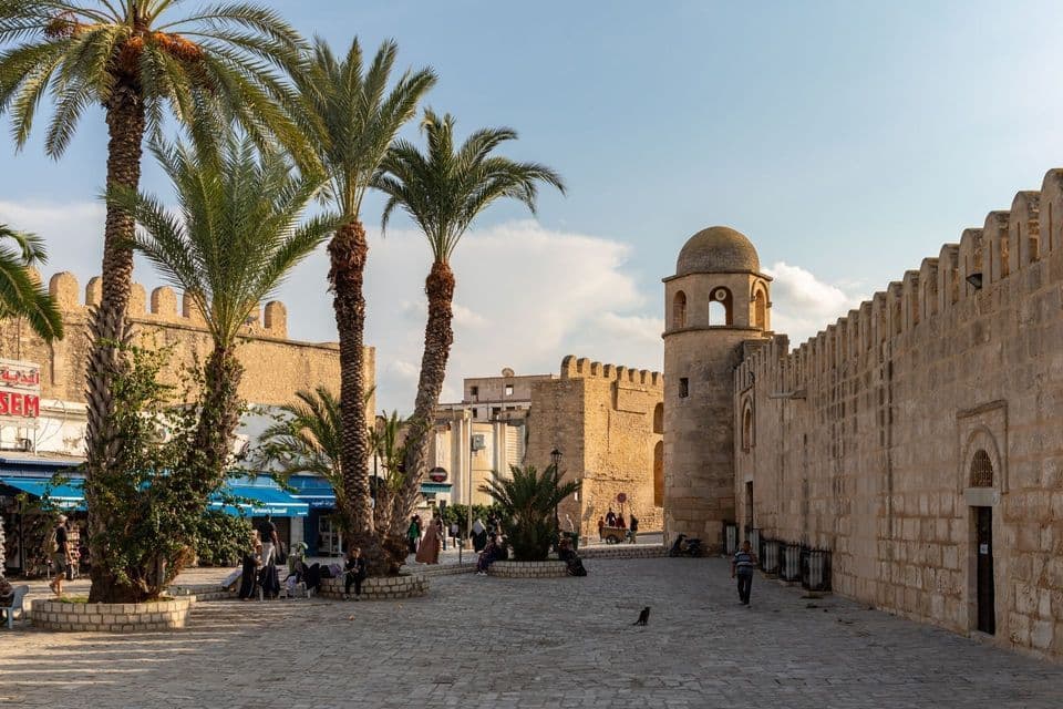 Ein sonnenbeschienener Kopfsteinpflasterplatz ist umgeben von alten Steinmauern, einem Kuppelturm und hohen Palmen, mit verstreuten Menschen.