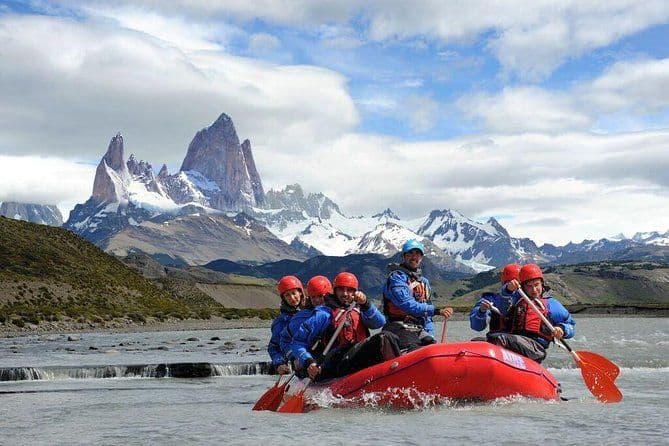 A WeRoad group trip in red helmets and life jackets rafting on a river in front of jagged, snow-covered mountains.