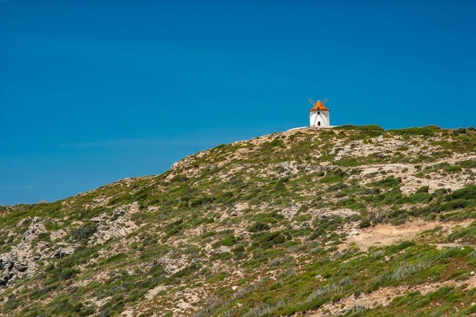 Eine weiße Windmühle mit rotem Dach steht auf einem felsigen, grünen Hügel unter einem weiten, klaren blauen Himmel.