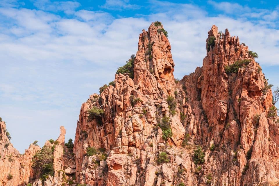 Zackige, rötliche Felsformationen mit kleinen grünen Bäumen stehen vor einem hellblauen Himmel mit weißen Wolken.