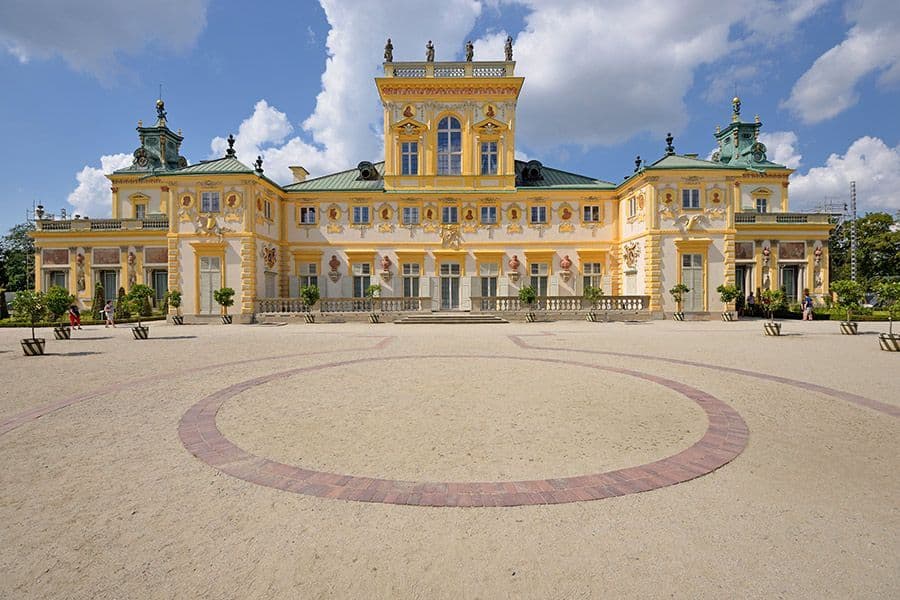 Un grand palais jaune orné, avec un toit vert, se dresse derrière une cour de sable, sous un ciel bleu parsemé de nuages blancs.