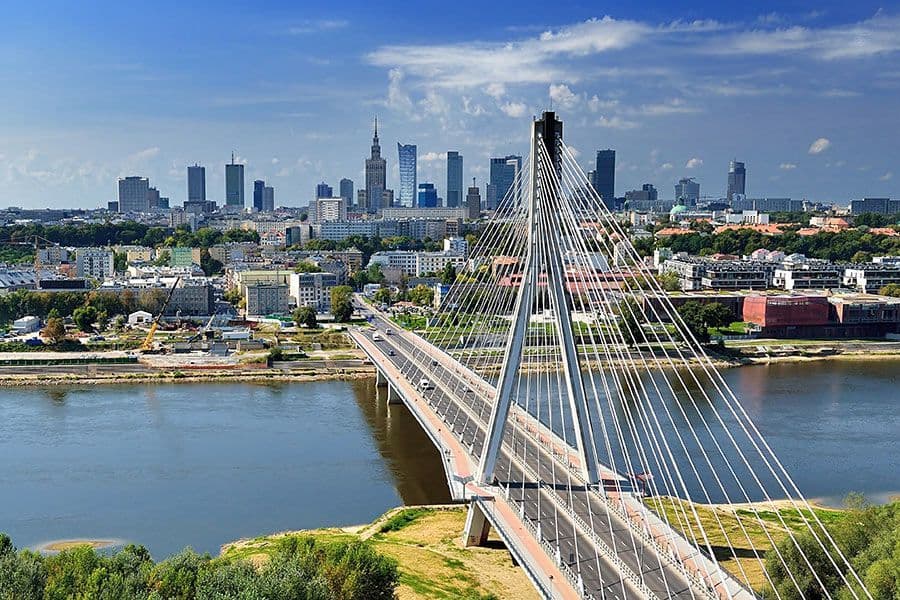 Un pont à haubans moderne enjambe une large rivière, menant vers un horizon urbain avec de nombreux gratte-ciel sous un ciel bleu partiellement nuageux.