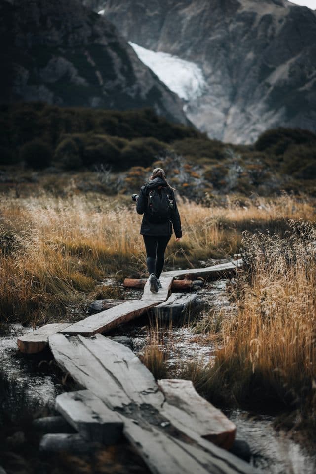 A hiker with a backpack walks on a wooden plank path through a field, with mountains and a glacier in the background.