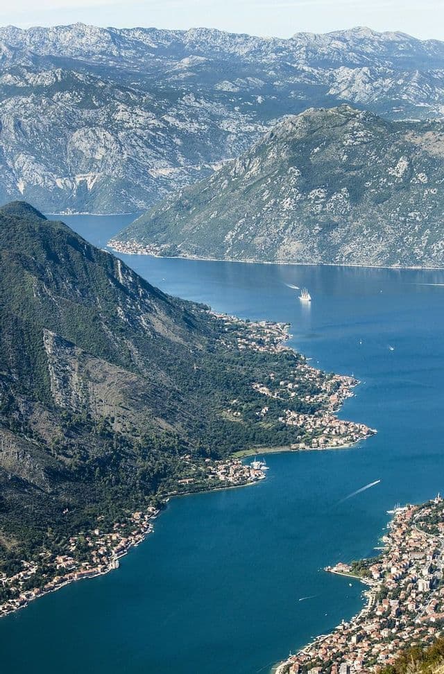 Vista aerea di una baia blu sinuosa, circondata da imponenti montagne frastagliate, con cittadine costiere adagiate lungo la riva.