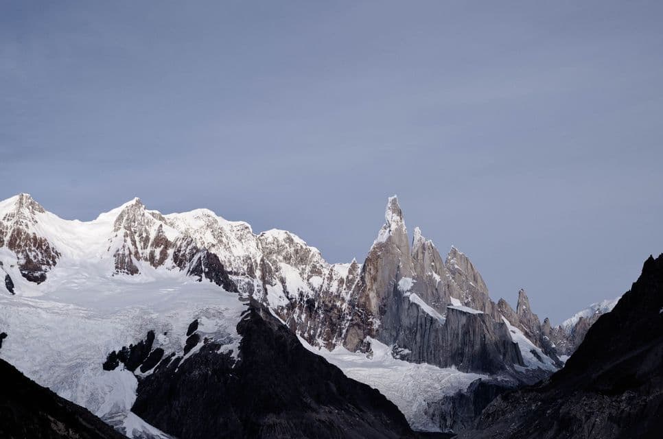 A rugged mountain range with sharp, snow-covered peaks and glaciers under a clear, pale sky.