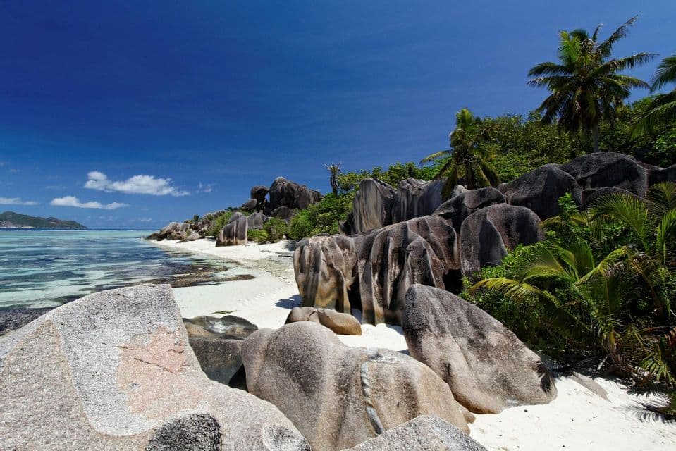 Große Granitfelsen ruhen auf einem weißen Sandstrand neben klarem, türkisfarbenem Wasser, mit üppigen Palmen unter einem tiefblauen Himmel.