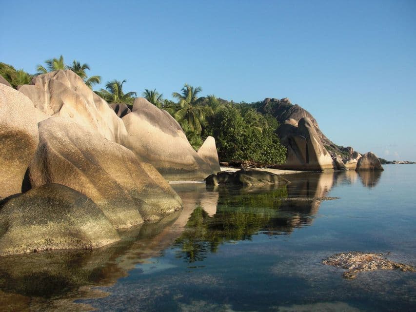 Große, glatte Granitfelsen liegen an einer tropischen Küste und spiegeln sich im ruhigen, klaren Wasser unter blauem Himmel wider.