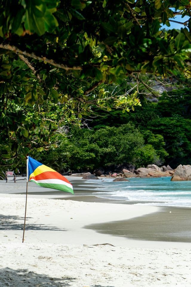 Die Flagge der Seychellen steht an einem weißen Sandstrand, mit türkisfarbenem Wasser und einem üppig grünen Wald im Hintergrund.