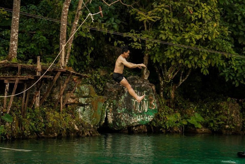 Un homme torse nu en short noir saute dans une rivière depuis une balançoire à corde dans une forêt dense et verdoyante.