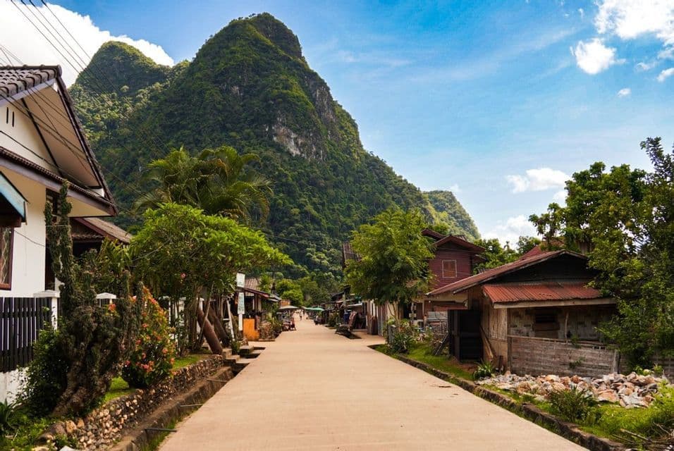 Une route pavée traverse un petit village avec des maisons et des arbres luxuriants, menant vers une grande montagne verte sous un ciel bleu.