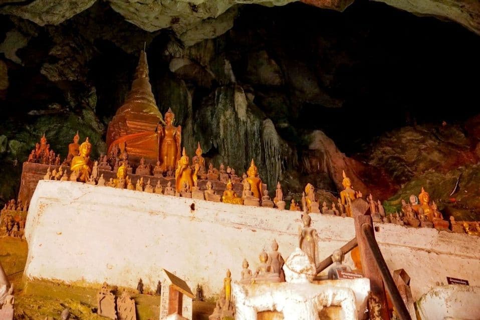 De nombreuses statues de Bouddha dorées et un stupa exposés sur une plateforme blanche à l'intérieur d'une grotte sombre et rocheuse.