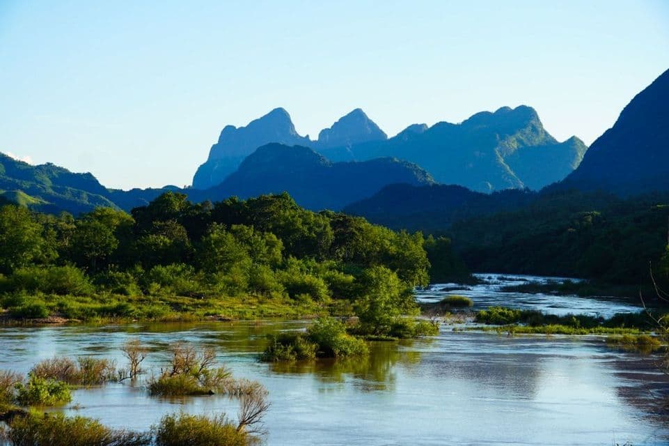 Une large rivière serpente à travers une vallée luxuriante et verte, avec des montagnes étagées se découpant sur le ciel bleu clair en arrière-plan.