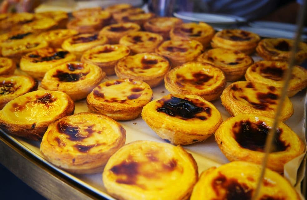 Un primo piano di numerosi pasteis de nata portoghesi con croste dorate e friabili e superfici caramellate, disposti su un vassoio.
