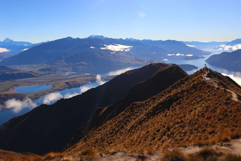 Un erboso crinale montano conduce a una vetta con due escursionisti che ammirano un vasto paesaggio di laghi e montagne sotto un cielo blu.