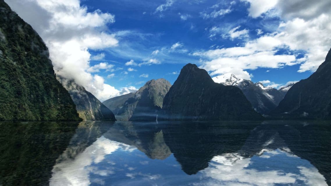 Steile grüne Berge und schneebedeckte Gipfel spiegeln sich im stillen Wasser eines Fjords unter einem bewölkten blauen Himmel.