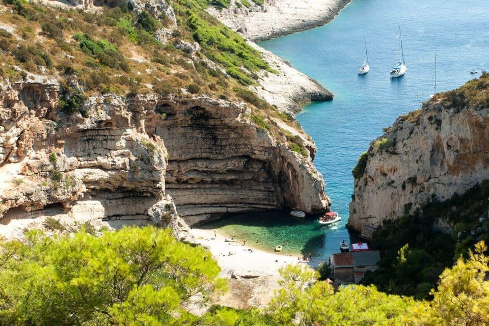 Eine Luftaufnahme eines abgelegenen Strandes, eingebettet zwischen hohen Felsklippen, mit Booten, die im türkisfarbenen Wasser ankern.
