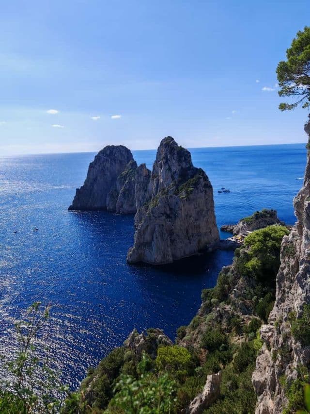 Una vista desde un acantilado alto muestra tres grandes farallones emergiendo de un mar azul brillante bajo un cielo despejado.
