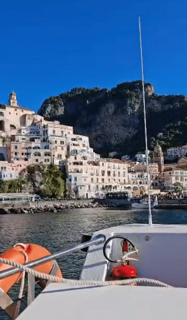 Una vista desde la proa de un barco mostrando un pueblo costero soleado con casas construidas en un acantilado empinado bajo un cielo azul claro.