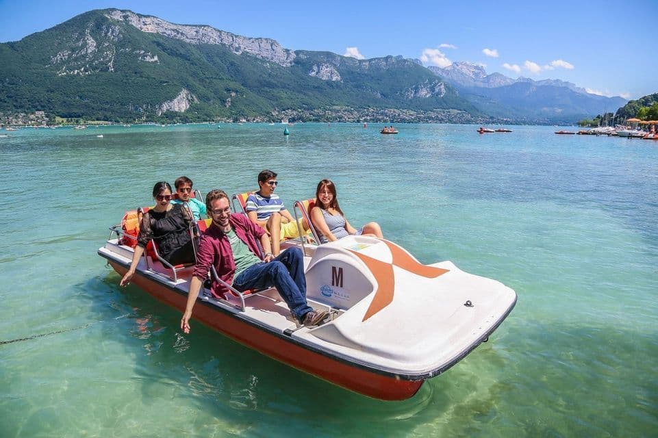 A WeRoad group trip of five people relaxing on a pedal boat on a clear lake with large, green mountains in the background.