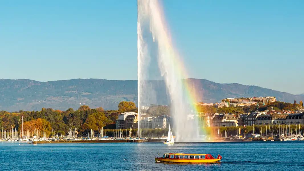 Ein großer Springbrunnen auf einem See erzeugt einen Regenbogen in seiner Gischt, während ein gelbes Boot vorbeifährt, mit einer Stadt und Bergen im Hintergrund.