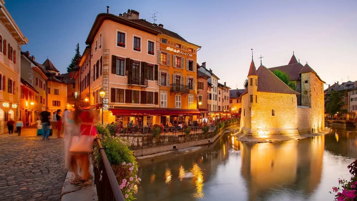 Un bâtiment historique en pierre se dresse dans un canal, entouré de restaurants et de boutiques illuminés le long d'une rue pavée au crépuscule.