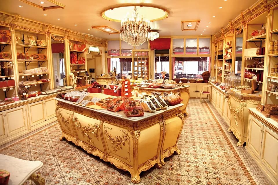 The ornate interior of a confectionary shop, with a crystal chandelier and large counters displaying various chocolates and sweets.