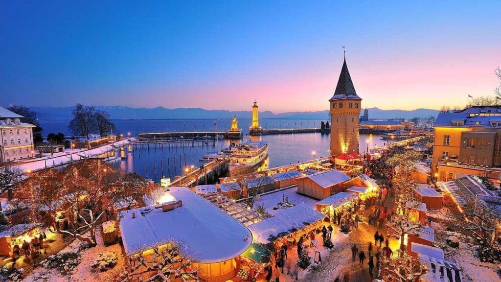 An elevated view of a snowy harbor town at dusk, featuring an illuminated Christmas market, a lighthouse, and a stone tower by the water.