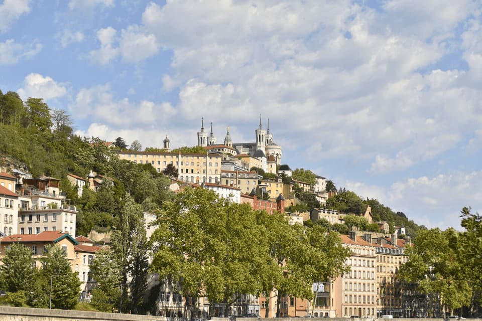 Une ville avec une basilique à son sommet est bâtie à flanc de colline verdoyante sous un ciel partiellement nuageux.