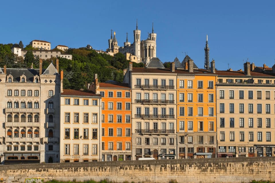 Une rangée de bâtiments colorés le long d'un front de mer, avec une grande basilique sur une colline verdoyante à l'arrière-plan sous un ciel bleu clair.
