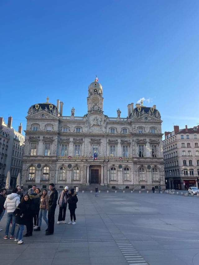Un groupe WeRoad se trouve sur une grande place ensoleillée, devant un bâtiment historique orné, sous un ciel bleu clair.