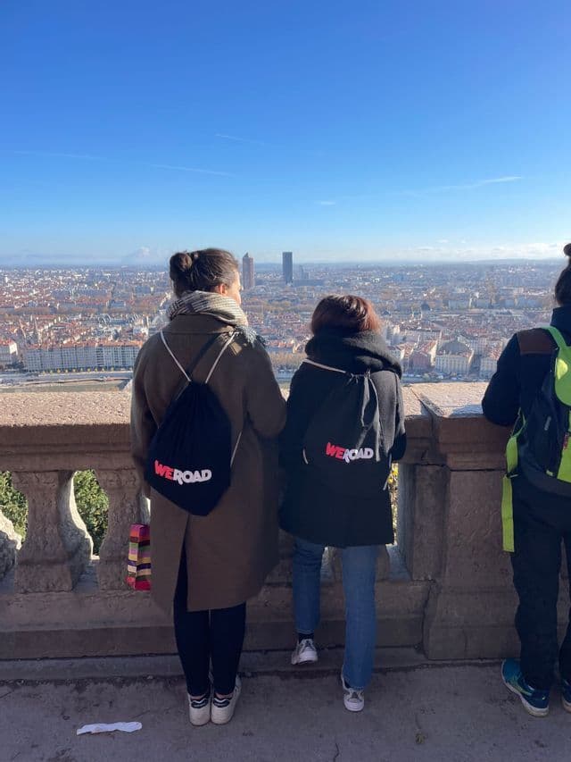 Trois membres d'un voyage de groupe WeRoad, vus de dos, admirant un paysage urbain depuis une balustrade en pierre, par une journée ensoleillée.