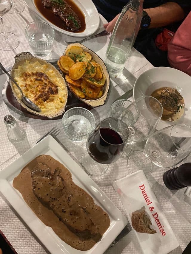 Vue plongeante sur une table de dîner richement garnie de divers plats : viande en sauce, gratin, frites et verre de vin rouge.