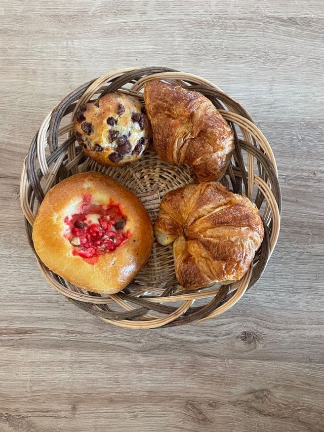 Vue plongeante d'un panier en osier contenant quatre pâtisseries, notamment des croissants et des muffins, posé sur une surface en bois clair.