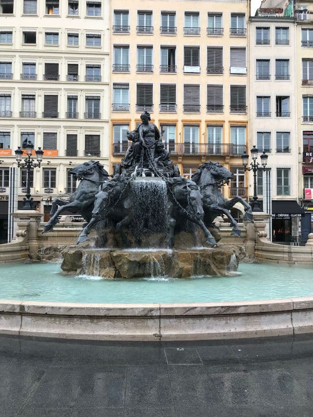 Une fontaine en bronze ornée, avec un char tiré par quatre chevaux, se dresse sur une place de ville devant de hauts bâtiments européens classiques.