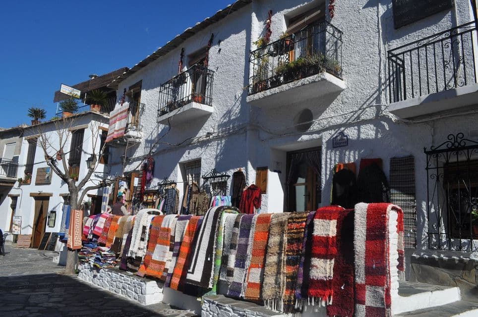 Alfombras tejidas coloridas y ropa expuestas a la venta en un muro bajo frente a un edificio encalado en un día soleado.