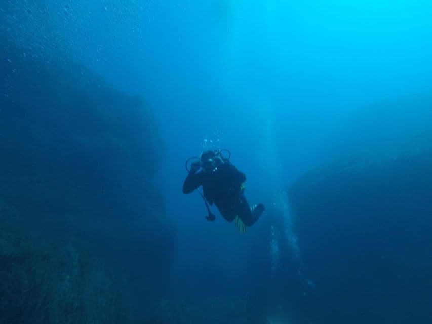 Un subacqueo in muta completa nuota attraverso un canyon sottomarino in acque blu profonde, circondato da formazioni rocciose.