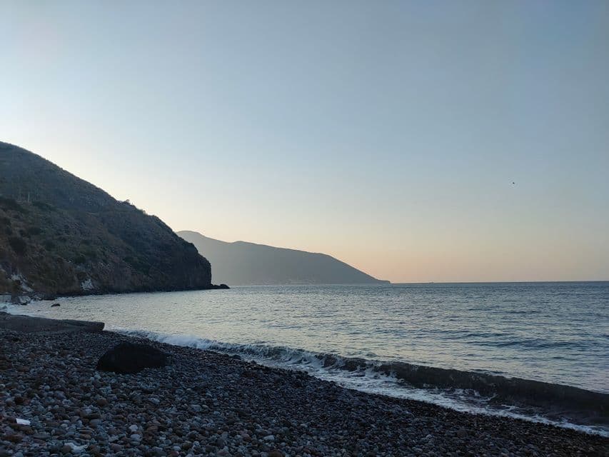 Una spiaggia di ciottoli con piccole onde che lambiscono la riva, accanto a una grande montagna, sotto un cielo limpido al tramonto.