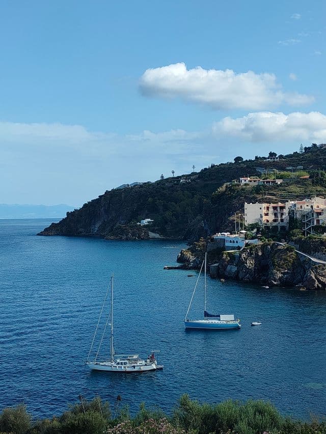 Due barche a vela ormeggiate in una tranquilla baia blu, accanto a una costa rocciosa e verdeggiante con case sulla scogliera.