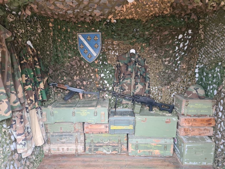 Militärische Darstellung mit zwei Gewehren auf Munitionskisten, Tarnuniformen, einem Helm und einem bosnischen Wappen vor einer Tarnnetzwand.