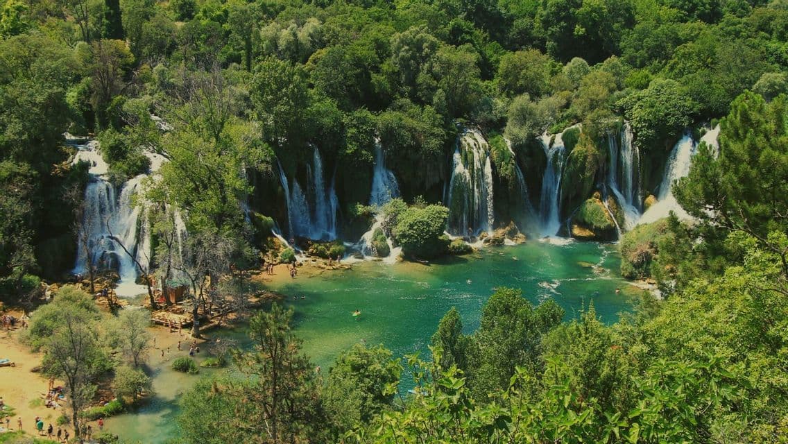 Ein Panoramablick auf mehrere Wasserfälle, die in einen türkisfarbenen Fluss stürzen, umgeben von einem üppig grünen Wald, mit schwimmenden Menschen darunter.
