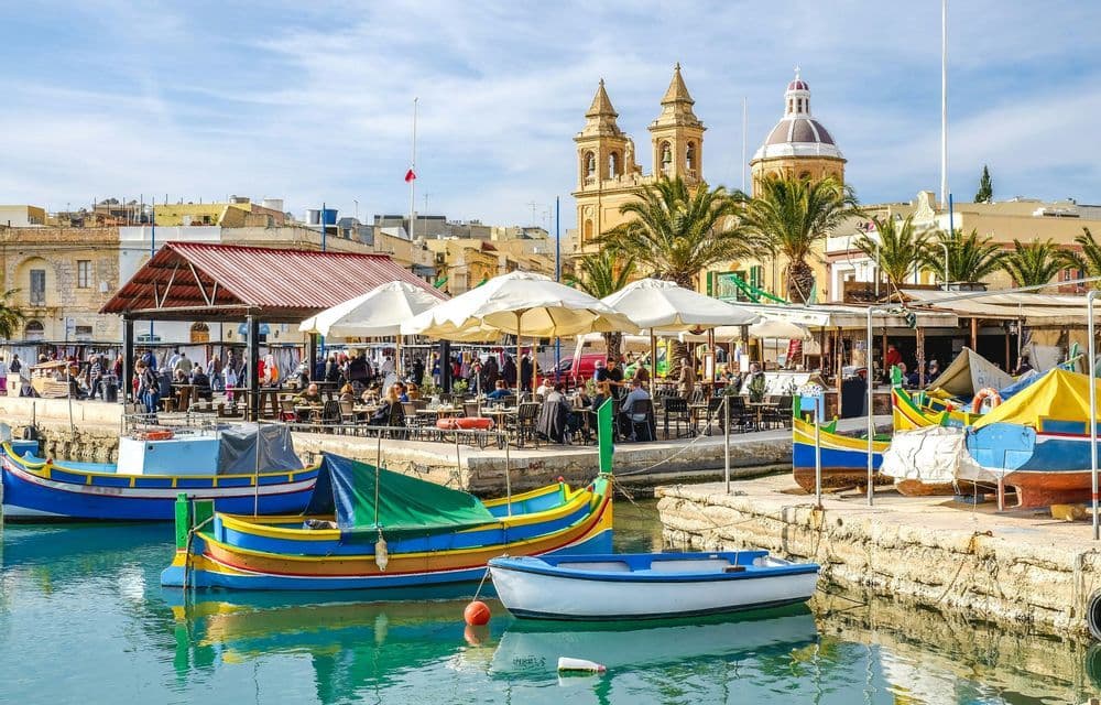 Des bateaux colorés sont amarrés dans un port, à côté d'une promenade en bord de mer où des gens dînent dans des cafés en plein air sous un ciel ensoleillé.