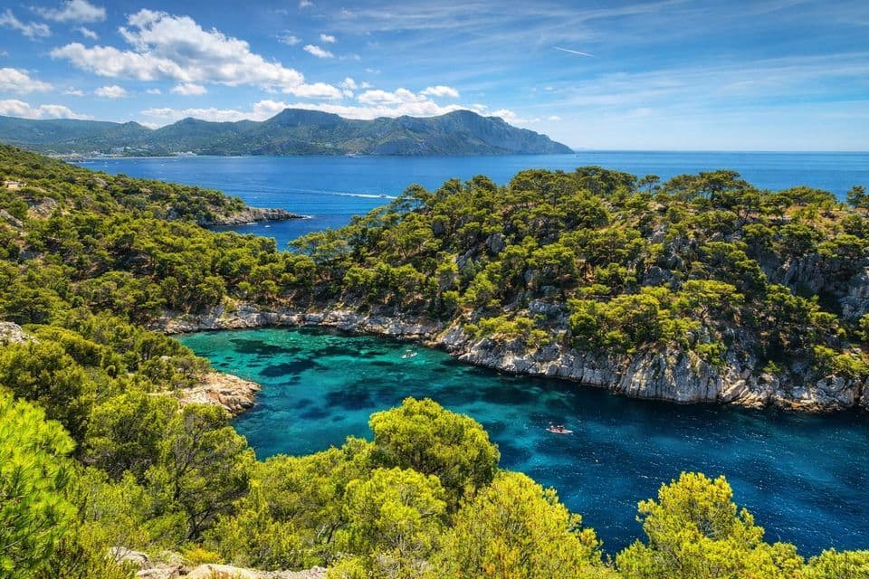 Una veduta dall'alto di una cala rocciosa con acqua turchese cristallina, circondata da colline ricoperte di pini, con montagne lontane oltre il mare.