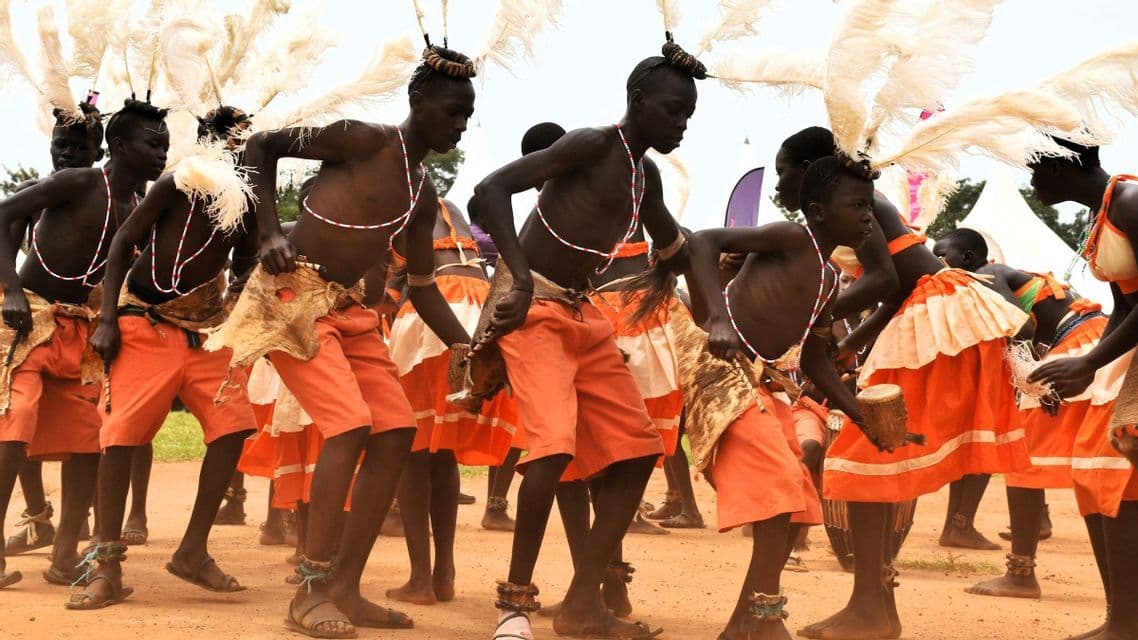 Eine Gruppe von Menschen in orangefarbenen traditionellen Gewändern und Federkopfschmuck führt einen kulturellen Tanz auf einem Erdboden auf.