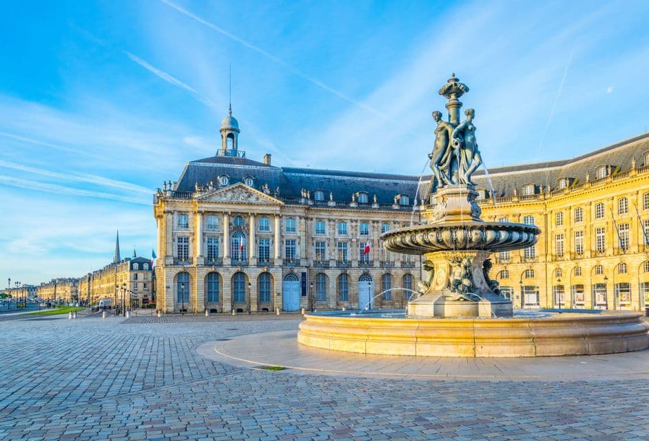 Une fontaine ornée de statues se trouve sur une place pavée ensoleillée, avec un grand bâtiment historique en arrière-plan.
