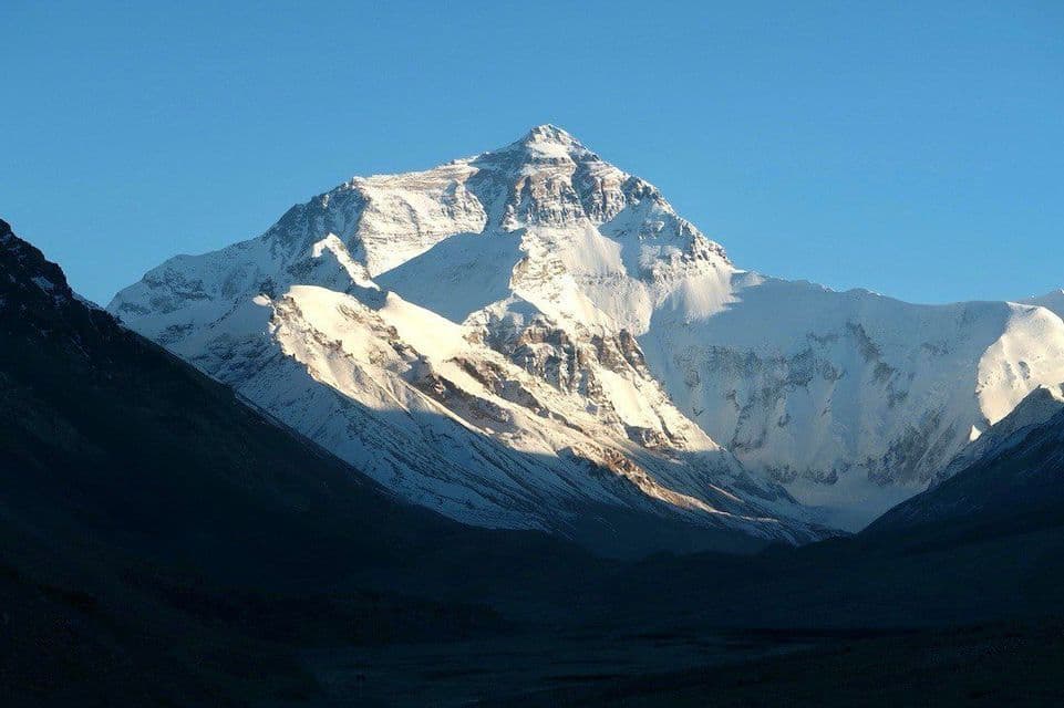 Una cima montuosa alta e innevata è illuminata dal sole contro un cielo blu limpido, sorgendo da una valle in ombra.