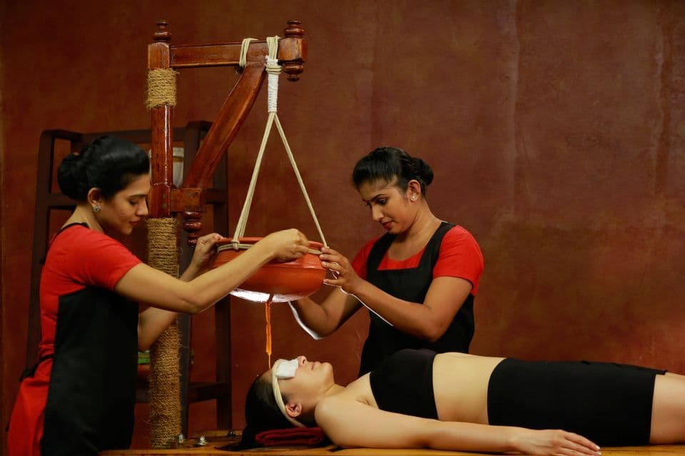 Una donna riceve un trattamento Shirodhara con olio, con i praticanti che versano olio da un vaso di argilla sospeso sulla sua fronte.