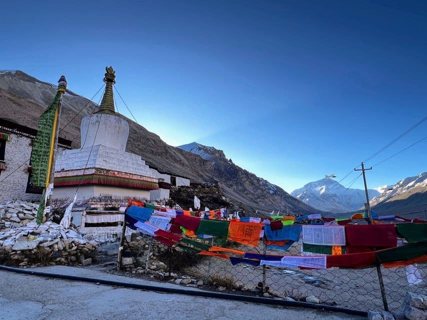 Uno stupa bianco sorge accanto a bandiere di preghiera colorate in una valle di montagna, con cime innevate visibili in lontananza sotto un cielo sereno.